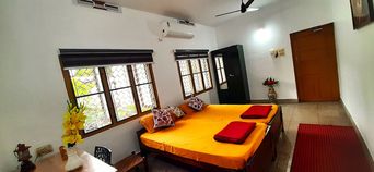 Bedroom with a double bed under bright orange sheets with red and patterned cushions, leaded-glass windows with roller blinds, a wall air conditioner and a red rug on a tiled floor.