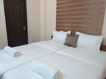 Bedroom with two white beds pushed together, rolled white towels at the foot, a brown cushion and a wood-slatted headboard, beside a dark wooden door.