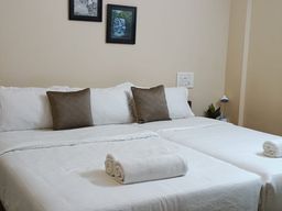 Bedroom with white bedding on two beds side by side, two brown cushions, two framed pictures on the wall and a small lamp on a bedside table.