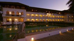 Long white colonial-style hotel with arched windows lit at dusk, reflected in a pool, with a stone dolphin sculpture in front.