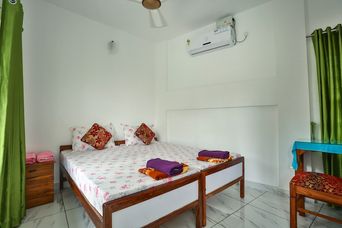 Bedroom with a double bed under a white floral sheet with red-patterned cushions, green curtains, and a wall-mounted air conditioner.