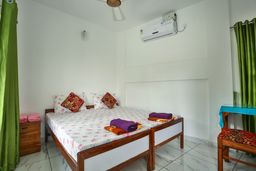 Bedroom with a double bed under a white floral sheet with red-patterned cushions, green curtains, and a wall-mounted air conditioner.