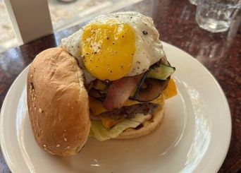 A burger on a white plate with the sesame bun set to one side: a beef patty with melted cheese and lettuce, grilled courgette and bacon stacked above it, and a fried egg with a bright yolk on top.