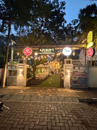 The entrance to a venue at dusk, framed by two white pillars under a metal archway strung with warm lights. A central sign displays a name above smaller text, flanked by circular illuminated signs on either side. Through the open gate, a path of artificial grass leads past white tables and chairs toward a building with an umbrella. A poster on the right pillar shows a cartoon coffee cup and mentions egg coffee. Trees with dense foliage rise above the structure against a darkening sky, and a paved brick walkway occupies the foreground.