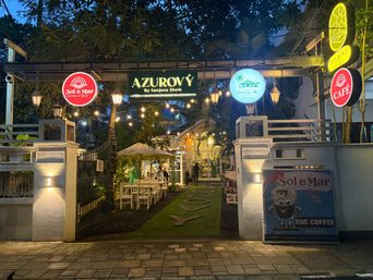 A gated entrance to an outdoor dining area at dusk, illuminated by hanging string lights and wall-mounted lamps. A central archway displays several illuminated signs, including a circular red sign, a green rectangular sign, a circular blue sign with a palm tree, and a vertical yellow sign with a circular red sign below it. To the right of the gate, a large standing board features an illustration of a coffee cup. Inside the gate, white tables and chairs are arranged under a white canopy on a grassy path lined with decorative cutouts. A person stands near the entrance, and trees and foliage surround the area.