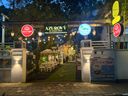 A gated entrance to an outdoor dining area at dusk, illuminated by hanging string lights and wall-mounted lamps. A central archway displays several illuminated signs, including a circular red sign, a green rectangular sign, a circular blue sign with a palm tree, and a vertical yellow sign with a circular red sign below it. To the right of the gate, a large standing board features an illustration of a coffee cup. Inside the gate, white tables and chairs are arranged under a white canopy on a grassy path lined with decorative cutouts. A person stands near the entrance, and trees and foliage surround the area.