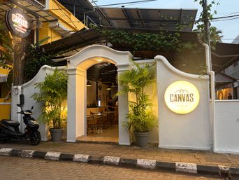A white arched entrance leads into a warmly lit interior space with wooden tables and chairs visible inside. Two potted palm trees flank the archway, one on each side. To the right of the entrance, a circular illuminated sign is mounted on the white wall. A black scooter is parked on the left side near a pole that holds another circular sign above it. The building has a yellow upper section with greenery growing along the roofline. A paved sidewalk with black and white curbs runs along the front.