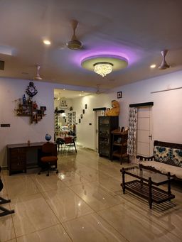 Living room with glossy beige floor tiles and ceiling fans, a purple-lit ceiling recess and chandelier. A wooden sofa with floral cushions, a glass-topped table, a desk and a bookshelf.