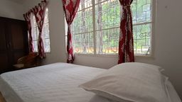 Bed with white linen in a bedroom, with red patterned curtains at a window behind an ornate iron grille, and a dark wooden wardrobe with a rattan armchair beside it.