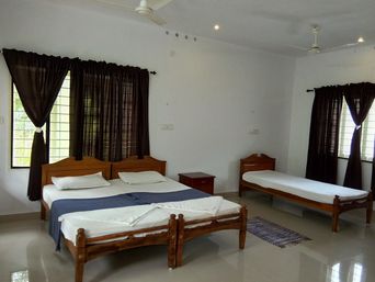 Bedroom with a wooden double bed and a single bed, both with white linen, dark brown curtains at tall windows, ceiling fans, a small wooden bedside table and a striped rug on a glossy floor.