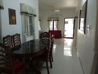 Dining area with an oval dark glass-topped table and wooden chairs with red cushions, lace-trimmed curtains and a framed picture. A glossy tiled hallway leads to a red sofa and a glass door.
