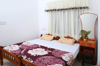 Bedroom with a carved wooden double bed under a rose-patterned maroon bedspread, cream sheer curtains with a tasselled valance, and a dressing table with an arched mirror.