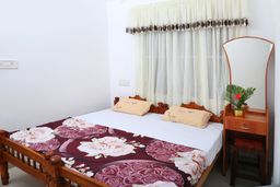 Bedroom with a carved wooden double bed under a rose-patterned maroon bedspread, cream sheer curtains with a tasselled valance, and a dressing table with an arched mirror.