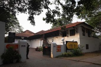 The white walls and red tiled roofs of Bastion Bungalow behind its gate, with signboards at the entrance and a large tree overhanging the drive.
