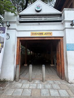 A white building with a triangular pediment and a clock at the top center. Below the clock is a long horizontal fixture with metal bars. A rectangular sign is mounted above a wide wooden doorway with double doors open inward. Three short, cylindrical stone bollards stand on the paved ground in front of the entrance. The interior is a long, covered corridor supported by wooden pillars, receding into shadow. A vertical banner is partially visible on the left side, and a blue informational board is mounted on the right wall. Trees with green leaves are visible above the roofline.