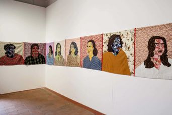 A long line of stitched fabric portraits hung at head height around two gallery walls, each face pieced from patterned cloth on a floral backing.