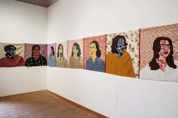 A long line of stitched fabric portraits hung at head height around two gallery walls, each face pieced from patterned cloth on a floral backing.