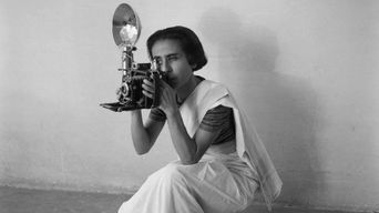 A black-and-white photograph of a woman in a sari crouching to shoot with a large press camera and flash reflector.