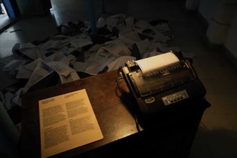 A dot-matrix printer on a wooden desk in a dim room, feeding a blank sheet, with hundreds of printed pages strewn across the floor behind it.