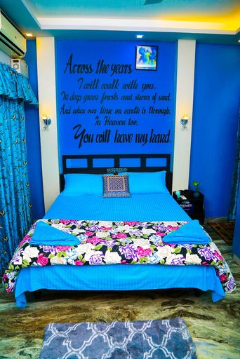 Bedroom with blue walls and blue bedding on a double bed with a floral throw, a black lettered wall decal above the headboard, blue curtains and a patterned rug.