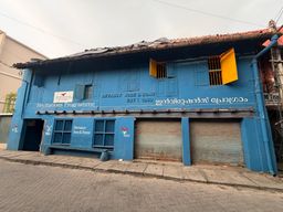 A two-story blue building with a weathered tiled roof stands on a paved street. The upper floor has several windows, some with yellow shutters open and others with black metal grilles. The ground floor features two closed metal roller shutters, a dark entrance on the left, and windows with black grilles beneath the upper windows. White text and logos are painted on the facade, including a sign near the entrance and another above the roller shutters. A vertical pipe runs along the right side of the building, and overhead wires cross the sky above.