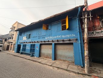 A long, single-story building painted bright blue stands on a paved street corner under an overcast sky. The building has a tiled roof and a row of closed metal roller shutters along the front. Several windows are visible, some with wooden frames and one with yellow shutters open. A vertical blue pipe runs down the right side of the building next to a utility pole covered in wires. To the left, another building with balconies is partially visible, and to the right, a neighboring structure has a red roof and a closed shutter. A sign is mounted on the blue wall above the shutters, and another sign is visible on the adjacent building to the right.