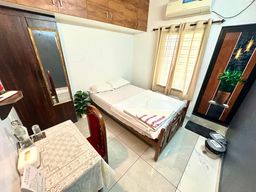 A small room with a single bed against a wall, a wooden wardrobe with a mirror on the left, a wall-mounted air conditioner above a curtained window, and a potted plant by the bed.