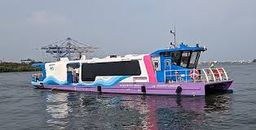 A blue, white and pink Kochi Water Metro boat on open water, with container cranes on the shore behind it.