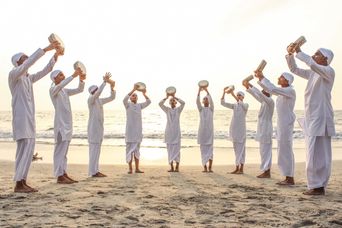 Ten young men in white kurtas and caps stand in a curve on a beach at sunset, each holding a duff, a round frame drum, above his head.