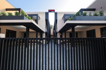 Black vertical-slat gate across a paved courtyard, with two-storey cream buildings on either side, dark flat-roofed overhangs and plants on the upper balconies.