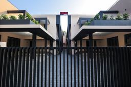 Black vertical-slat gate across a paved courtyard, with two-storey cream buildings on either side, dark flat-roofed overhangs and plants on the upper balconies.