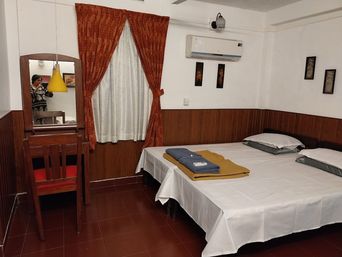 Bedroom with wood-panelled lower walls and dark red tiles, two beds with white sheets and folded towels, red curtains, a wall air conditioner and a dressing table with a yellow lamp.