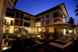 Three-storey hotel at dusk around a dark pool, with sun loungers and parasols on the deck and lit rooms behind.