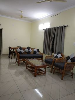 Living room with a cane sofa and two cane armchairs with dark cushions, a glass-top wooden coffee table, grey curtains, and pale yellow walls.
