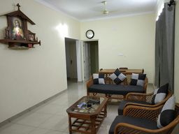 Living room with pale yellow walls and glossy tiled floor, cane sofa and armchairs with dark grey cushions, a glass-topped wooden coffee table, a wall clock and a framed picture on a shelf.