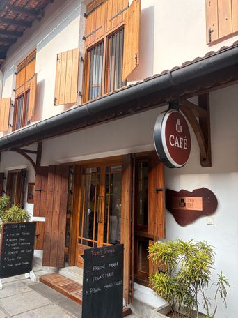 A two-story building with white walls and wooden shutters on the upper windows. A round hanging sign hangs above a wooden entrance with glass doors. To the right of the entrance, a wooden plaque is mounted on the wall. Two black chalkboards stand on the paved sidewalk in front. Potted green plants are placed near the entrance and along the side of the building.