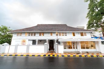 White two-storey colonial-style building with a tiled sloping roof and rows of tall windows, a small arched doorway and a paved forecourt edged in yellow and black, beside a wet road.
