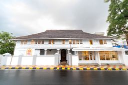 White two-storey colonial-style building with a tiled sloping roof and rows of tall windows, a small arched doorway and a paved forecourt edged in yellow and black, beside a wet road.
