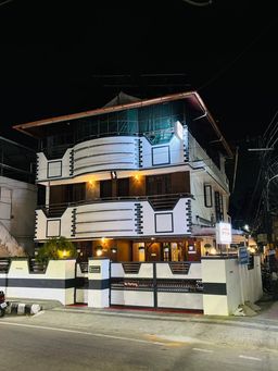 Three-storey white building at night with curved balconies and lit windows, behind a low wall with black stripes and a gate with a no-parking sign, beside a road.