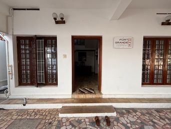 Entrance of a white building with two wooden-framed windows and an open wooden doorway, flanked by wall lamps, with a sign beside the door and a tiled forecourt in front.