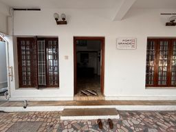 Entrance of a white building with two wooden-framed windows and an open wooden doorway, flanked by wall lamps, with a sign beside the door and a tiled forecourt in front.