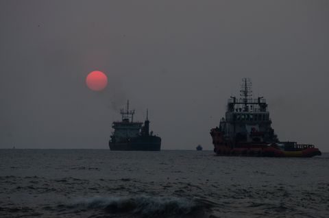 A red sun low in a hazy grey sky over the sea off Fort Kochi Beach, with two ships at anchor and a small wave breaking in the foreground.