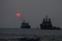 A red sun low in a hazy grey sky over the sea off Fort Kochi Beach, with two ships at anchor and a small wave breaking in the foreground.