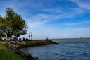 The rock-lined shore at Fort Kochi Beach on a bright day: a large tree and food stalls on the left, two lamp posts at the water’s edge, a few people on the rocks and a ship far out at sea.
