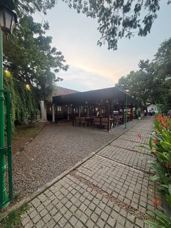 A covered outdoor dining area with tables and chairs sits under a dark roof, surrounded by trees and greenery. A paved walkway runs alongside a gravel path, bordered by a green metal post on the left and flowering plants on the right. The sky above is overcast with soft light filtering through the tree canopy.
