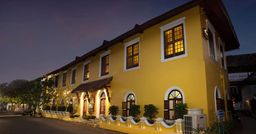 A yellow two-storey colonial-style building at dusk, its windows lit, with an arched entrance under a small canopy and potted plants along a white balustrade by the street.