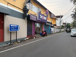 A street scene shows a row of shops with closed metal shutters under a yellow building, one with a purple sign and another with a pink logo. A blue directional sign on two posts stands on the sidewalk near a red brick wall. A red scooter is parked in front of one shop, and a white car and a black auto-rickshaw are on the road ahead. Overhead wires run across the sky above the street.