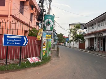 A street scene shows a blue directional sign with white text and an arrow pointing right, mounted on a black post beside a red metal fence. Behind the fence is a peach-colored building with dark windows. A utility pole stands nearby, covered with several posters and signs, including a large green one with a circular graphic. To the right, a paved road curves gently into the distance, lined with buildings, trees, and parked motorcycles. A white sign with red lettering lies on the grass at the base of the fence.