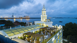 Rooftop terrace at dusk with wooden pergolas, laid tables and chairs, and a glass-roofed section beside a white clock tower above a harbour.