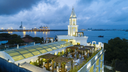 Rooftop terrace at dusk with wooden pergolas, laid tables and chairs, and a glass-roofed section beside a white clock tower above a harbour.